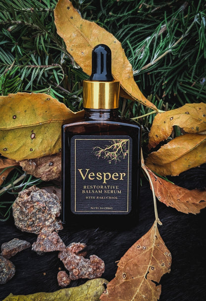 Vesper Restorative Balm Serum bottle with dropper on a dark surface with leaves and stones. This serum includes tree saps and resins as well as bakuchiol, pomegranate seed, black cumin seed, carrot seed, prickly pear seed, camellia seed, and jojoba oil. anti-aging and acne friendly natural skin serum. 